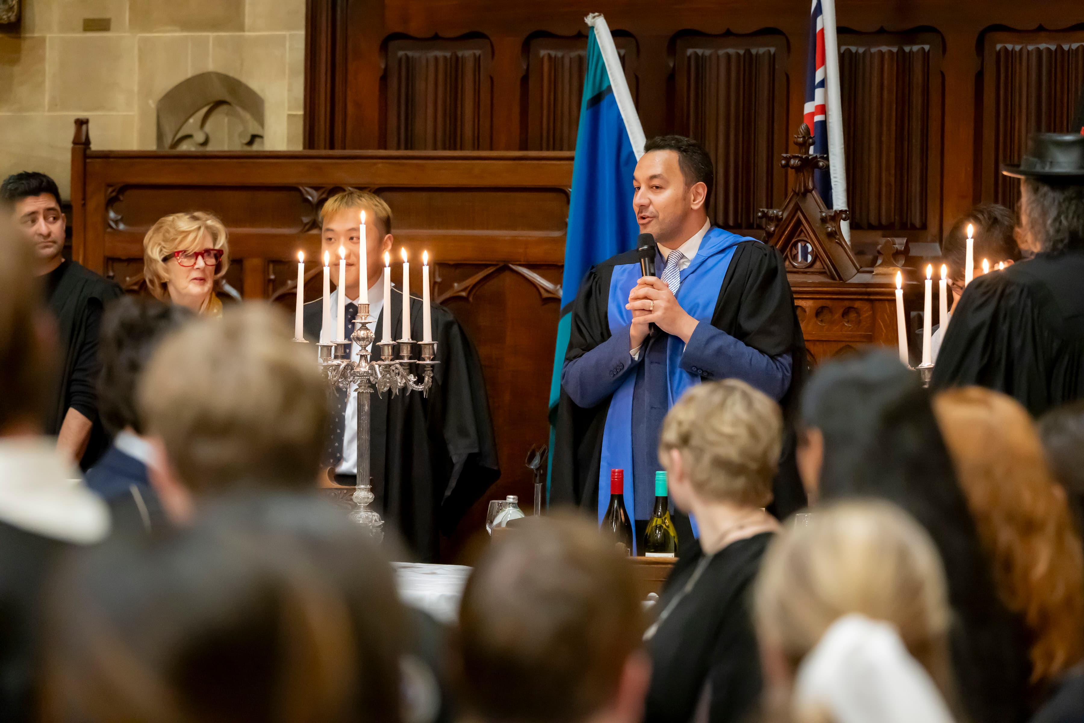 Dr Areti (Ari) Metuamate dressed in an academic gown with microphone the Ormond College Dining Hall.